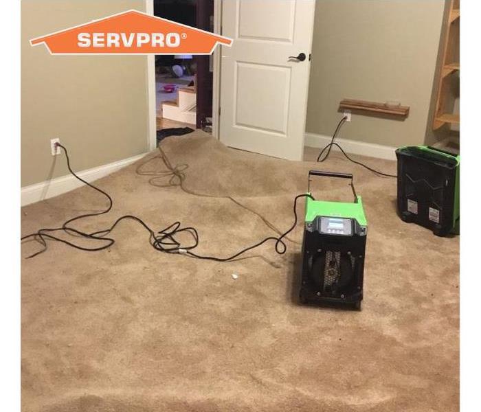 Two pieces of SERVPRO green quipment drying out brown carpet in a water damaged home. Orange SERVPRO house in corner